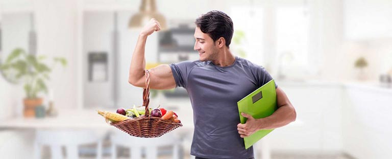 man flexing his bicep with a basket of fruit and vegetables hanging from his arm.